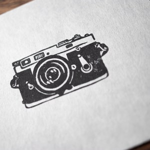 Op de afbeelding: Zwart-witte illustratie van een vintage camera met een lens en een filmterugspoelknop.