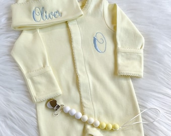 May include: A pale yellow baby outfit set, including a footed onesie, a matching cap, and a pacifier clip. The cap is embroidered with the name "Oliver" in light blue. The onesie features a large embroidered "O" and scalloped trim.