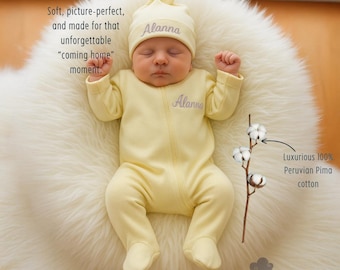 May include: A newborn baby wearing a pale yellow footed sleeper and matching cap, both embroidered with the name "Alanna". The baby is lying on a white, fluffy surface. The image includes text that says "Luxurious 100% Peruvian Pima cotton".