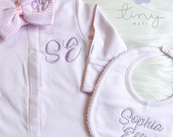 Puede incluir: Un conjunto de ropa para bebé de color rosa con un gorro, un lazo, un mono y un babero. El mono tiene las iniciales "SE" bordadas en la parte delantera. El babero tiene el nombre "Sophia Elliot" bordado. El conjunto es de la marca "tiny mati".