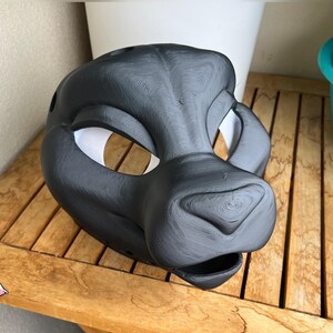 Puede incluir: Una máscara de animal negra mate impresa en 3D con un hocico inspirado en felinos, crestas de cejas prominentes y grandes aberturas para los ojos con forro interior blanco. La superficie muestra líneas de capa horizontales visibles.