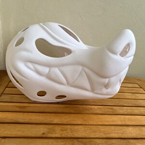 Op de afbeelding: Witte, ongeverfde gipsen masker van een grommend wezen met grote tanden en gaten voor ogen en neus.