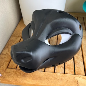 May include: A matte black animal-inspired mask featuring a sculpted snout, prominent brow ridges, and large eye openings with white inner linings. The mask includes small circular ventilation holes along the sides and a defined jawline structure.