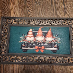 May include: A rectangular doormat with a decorative border. The center features a teal background with a black truck carrying three gnomes with red and white hats. The text "MERRY CHRISTMAS" is displayed.