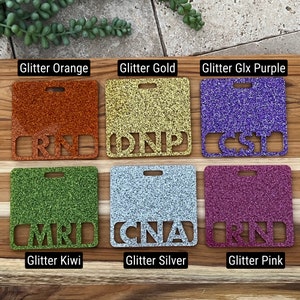 May include: Six square-shaped name tags with different glitter colors and text. The colors are orange, gold, purple, green, silver, and pink. The text on the tags reads "RN", "DNP", "CST", "MRT", "CNA", and "RN".