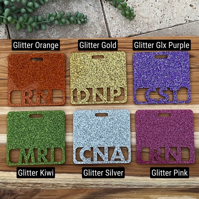 May include: Six square-shaped name tags with different glitter colors and text. The colors are orange, gold, purple, green, silver, and pink. The text on the tags reads "RN", "DNP", "CST", "MRT", "CNA", and "RN".