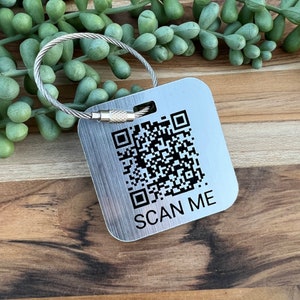 May include: A square, brushed metal luggage tag with a QR code and the words "SCAN ME" printed in black. The tag is attached to a silver-colored, flexible metal loop.