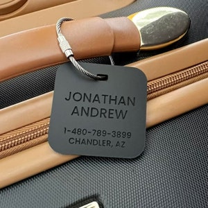 May include: A black luggage tag with the name "JONATHAN ANDREW" and a phone number and address. The tag is attached to a suitcase handle with a metal wire. The suitcase has a brown leather handle and a gold-colored accent.