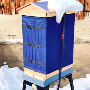 May include: A wooden beehive with a blue insulated cover and a black strap is on a wooden stand with black legs. The beehive is covered in snow.