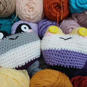 May include: Two handmade crochet frog-shaped items, one in grey, white, and purple, the other in white, purple, and yellow. The frogs have button eyes and are surrounded by colourful yarn balls in various shades.