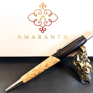 May include: A wooden pen with a silver clip and a cork-like barrel. The pen is resting on a small rock with gold-colored crystals. The pen is on a white surface with the word "AMARANTH" in gold lettering.