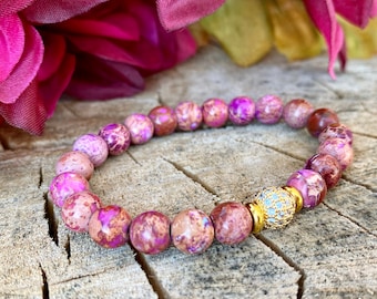 May include: A pink and brown beaded bracelet with a gold and clear crystal accent. The bracelet is on a wooden surface.