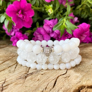 May include: Three white beaded bracelets of different textures and sizes are stacked. One bracelet features a silver bead with small, sparkling embellishments. The background includes pink flowers and green foliage, set on a wooden surface.