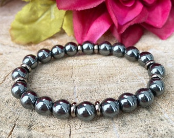 Puede incluir: Un brazalete de cuentas de hematites negras con detalles plateados. El brazalete está hecho de cuentas redondas y se muestra sobre una superficie de madera.