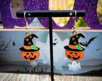 May include: A pair of gold-tone earrings featuring a pumpkin with a witch's hat. The pumpkin is orange with a black hat and green trim. The earrings are hanging from a black stand.
