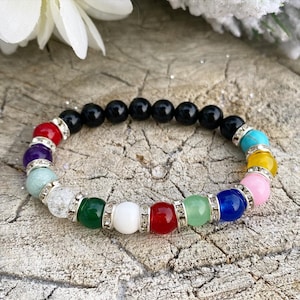 Puede incluir: Un brazalete de cuentas de colores con cuentas de ónix negro, cuentas de cristal transparente y cuentas de piedras preciosas de varios colores. El brazalete está sobre una superficie de madera.