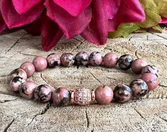 Puede incluir: Un brazalete de piedras preciosas rosa y negro con una cuenta de acento de oro rosa. El brazalete está hecho de cuentas redondas y tiene una cuenta de acento brillante y facetada.