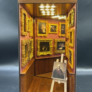 May include: Miniature art gallery diorama with a wooden frame. The interior features framed paintings on red walls, a wooden floor, and a small easel with a portrait. The gallery is illuminated by small lights.