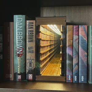 Puede incluir: Una estantería de madera con libros, incluyendo títulos de Dan Brown y Harry Potter. Una pieza decorativa en forma de entrada de biblioteca crea una ilusión de profundidad y luz entre los libros.