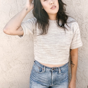 May include: A woman wearing a light gray cropped sweater with a light brown and white pattern and blue jeans.