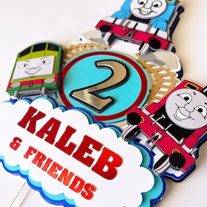 Könnte beinhalten: Ein Kuchentopper zum Thema Thomas die Lokomotive mit einer blauen, roten und grünen Lokomotive, einer silbernen Zahl 2 und einer weißen Wolke mit dem Text "Kaleb & Friends" in roten Buchstaben.