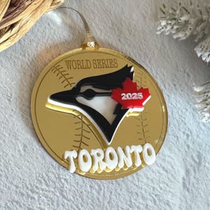 Puede incluir: Adorno redondo dorado con el texto "WORLD SERIES" y "TORONTO". El adorno presenta un logotipo de arrendajo azul con una hoja de arce roja y el año "2025". El fondo tiene costuras de béisbol.