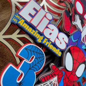Pode incluir: Uma decora&ccedil;&atilde;o de festa de Spiderman vermelha e azul com fundo branco. A decora&ccedil;&atilde;o apresenta o nome "Elias and his Amazing Friends" e o n&uacute;mero "3" com um design de teia de aranha.