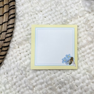 May include: A yellow note card with a blue border and a blue forget-me-not flower with a bee on it.