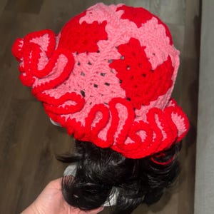 May include: A handmade crochet hat in a unique design. The hat features a pink and red color scheme with a wavy red trim. The hat is designed with a floral pattern and is a stylish accessory.