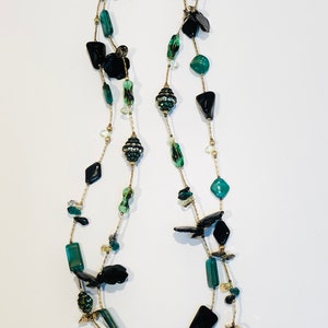 May include: A long, multi-strand necklace with black and green beads. The necklace features a variety of shapes and sizes of beads, including some that are faceted. The necklace is strung on a gold chain.