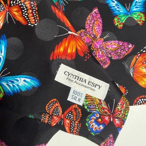 May include: A black silk scarf featuring a pattern of colorful butterflies in shades of orange, blue, pink, and green, with subtle dark gray polka dots. A white fabric label reads CYNTHIA ESPY Fine Accessories and 100% SILK in black text.