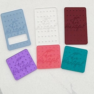 May include: A collection of colorful, rectangular acrylic charms. The charms are in shades of blue, clear, burgundy, purple, pink, and teal. Each charm has the words "you are beautiful" engraved on it.