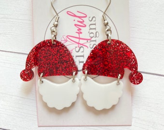 May include: A pair of festive Santa Claus earrings. Each earring features a red glitter Santa hat with a white beard. The earrings are suspended from silver-colored fish hook ear wires. The earrings are on a white card with the text "Amil Designs".