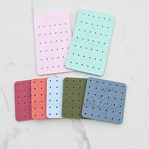 May include: A collection of colorful, rectangular tags with rounded corners. The tags are in shades of pink, coral, light blue, green, and blue. Some tags have the text "you are beautiful" and "Gisela" in cursive script, along with a pattern of small dots.
