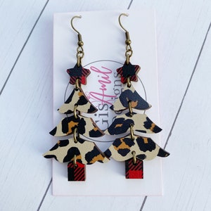 May include: Christmas tree-shaped earrings with a mix of patterns. The earrings feature a star at the top, followed by layers of leopard print and red and black plaid. The earrings hang from bronze-colored hooks.