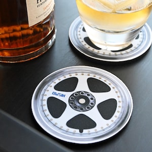 May include: Two white coasters with a black and silver rim and a black and white image of a car wheel with the word "NISSAN" printed on it.