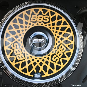 Puede incluir: Un tocadiscos negro con una aguja dorada y una alfombrilla antideslizante amarilla y negra con la palabra "BBS" impresa.