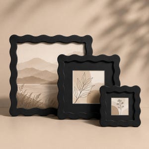 Op de afbeelding: Drie vierkante fotolijsten met een matzwarte afwerking en een golvende, geschulpte rand. De lijsten bevatten prints in beige en bruine tinten, waaronder een landschap met bergen, een lijntekening van bladeren en een kleine bloementak.