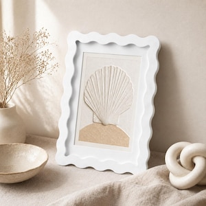 Può includere: Una cornice rettangolare bianca con un bordo ondulato e smerlato. All'interno della cornice si trova un'opera d'arte strutturata con un rilievo di conchiglia bianco sporco centrato sopra una forma semicircolare color marrone chiaro.