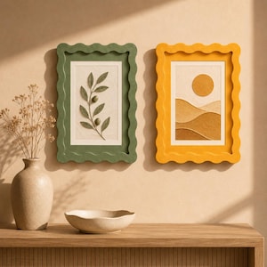 May include: Two rectangular wall frames with wavy edges, one in olive green featuring an embossed olive branch and one in mustard yellow displaying an embossed sun over rolling hills, alongside a beige vase with dried flowers and a bowl.