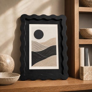 Peut inclure: Un cadre photo rectangulaire doté d'une bordure noire ondulée et festonnée. À l'intérieur se trouve une impression abstraite représentant un cercle noir uni au-dessus de lignes ondulées, texturées et superposées dans des tons beige, ocre et noir.