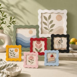 Peut inclure: Une collection de sept décorations murales carrées avec des cadres ondulés de différentes couleurs, présentant des motifs brodés de plantes, de paysages, d'une fleur et d'une part de gâteau aux fraises sur un fond en tissu clair.