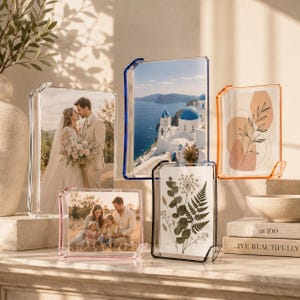 May include: A collection of five transparent acrylic picture frames in various colors including clear, blue, orange, pink, and black. The frames hold photographs of a wedding couple, a coastal landscape, a family, and botanical illustrations.