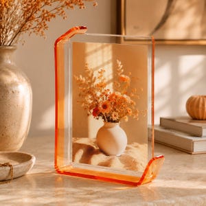 Könnte beinhalten: Ein rechteckiger, transparenter Acryl-Bilderrahmen mit leuchtend orangefarbenen Kanten. Der Rahmen verfügt über einen U-förmigen Sockel und einen Akzent an der oberen Ecke und enthält einen Druck mit einer kleinen, runden, cremefarbenen Vase voller orangefarbener und gelber Blumen.