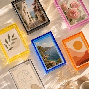Pode incluir: Uma coleção de seis molduras retangulares de acrílico em várias cores, incluindo preto, rosa, amarelo, azul e laranja. Cada moldura contém uma impressão diferente, como uma rua costeira, peônias rosas, uma ilustração de folha e uma vista do mar.