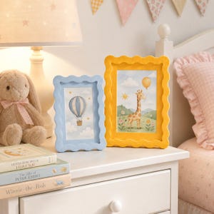 May include: Two rectangular picture frames with wavy edges, one light blue and one yellow, displaying illustrations of a hot air balloon and a giraffe with a balloon. The frames sit on a white surface next to a plush rabbit and stacked books.