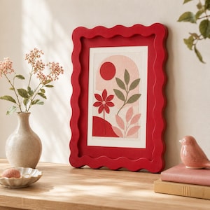 May include: A rectangular picture frame with a wavy, scalloped edge in a solid red finish. The frame contains a print featuring a red sun, a red flower, and green and pink foliage set against a cream-coloured background with an arch shape.