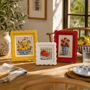 May include: Three scalloped picture frames in yellow, white, and red, each displaying a different botanical or food-themed print. The yellow frame holds a lemon illustration, the white frame shows tomatoes, and the red frame features red flowers.