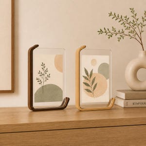 K&ouml;nnte beinhalten: Zwei rechteckige Bilderrahmen mit transparenten Acrylplatten und Holzsockeln. Ein Rahmen hat ein dunkelbraunes Holzfinish, der andere ein helles Naturholzfinish. Jeder Rahmen enth&auml;lt einen Druck mit botanischen Illustrationen und geometrischen Formen.