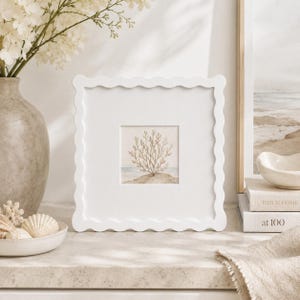 May include: A square white picture frame with a scalloped edge featuring a centred print of a tan coral-like plant on a sandy beach against a light blue horizon. The frame is matte white and has a clean, smooth finish surrounding the artwork.